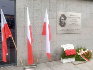 wieńce pod tablicą pamiatkową Danuty Siedzikówny "Inki",  trzy flagi biało-czerwone w stojaku obok tablicy